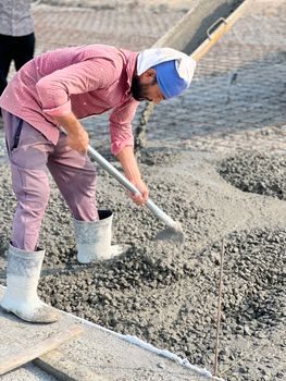 ouvrier étalant au râteau un béton frais dans lequel on distingue de fines fibres métalliques grises mélangées à la pâte, lumière de chantier en extérieur
